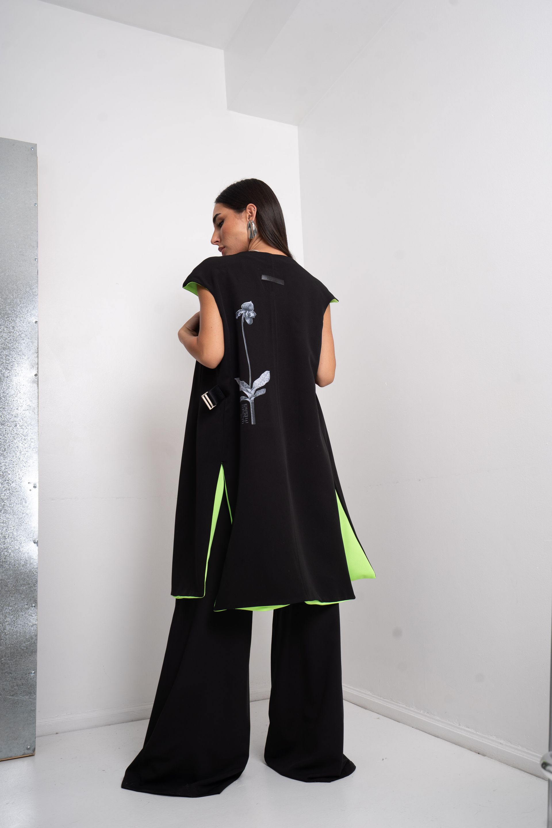 Mujer con chaqueta negra sin mangas, top verde y pantalones, de pie con la mano en la cabeza.