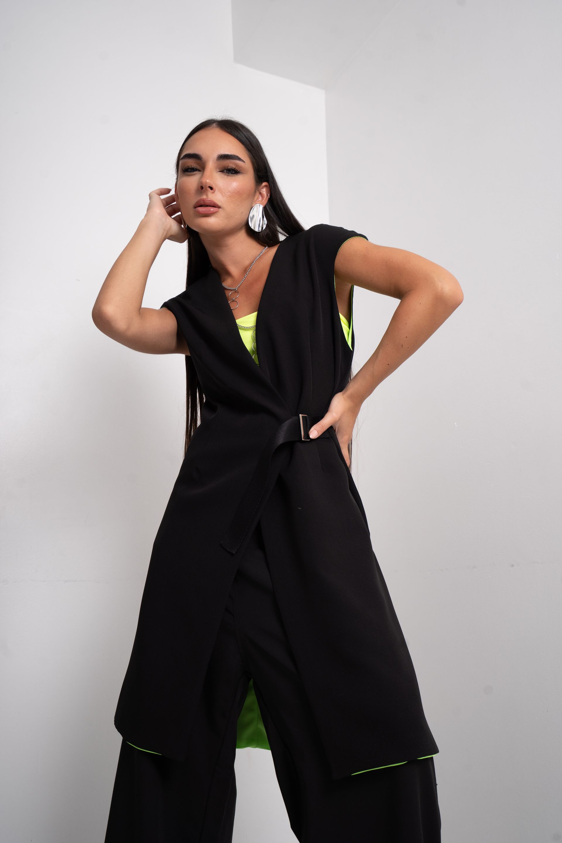 Mujer con chaqueta negra sin mangas, top verde y pantalones, de pie con la mano en la cabeza.