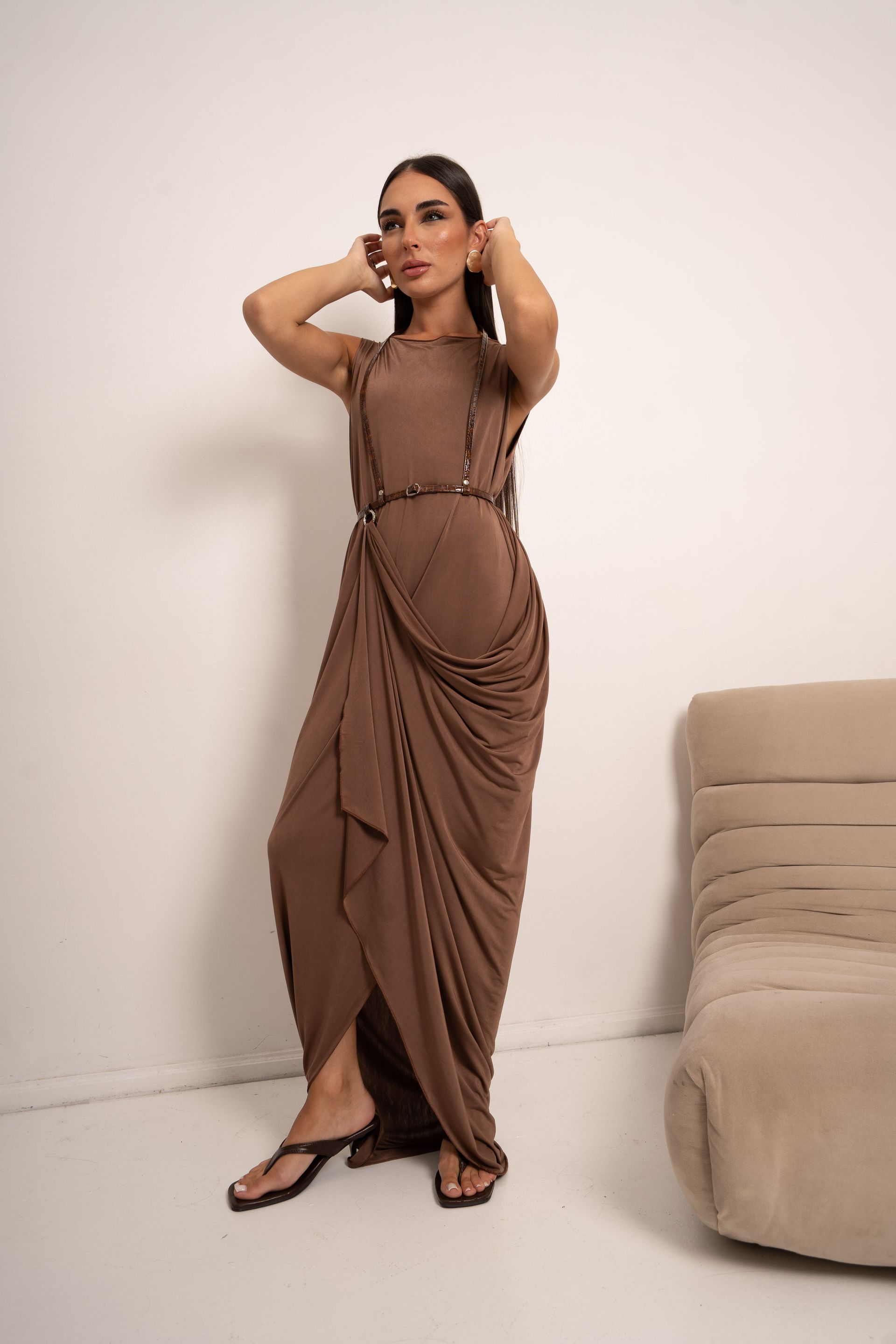 Mujer con un vestido marrón drapeado, con las manos cerca de la cabeza, de pie cerca de una silla beige.
