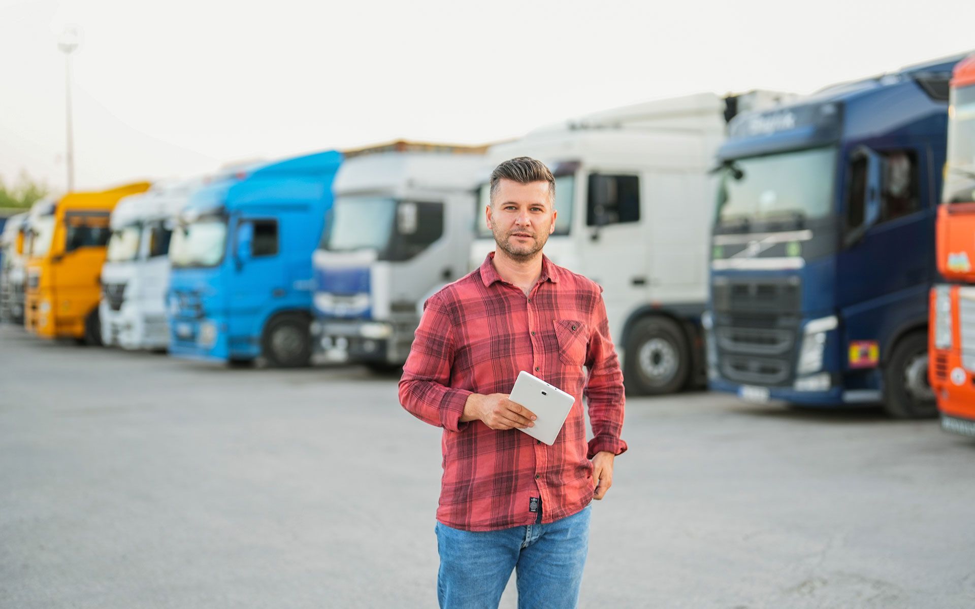 A man is standing in front of a row of semi trucks holding a tablet.