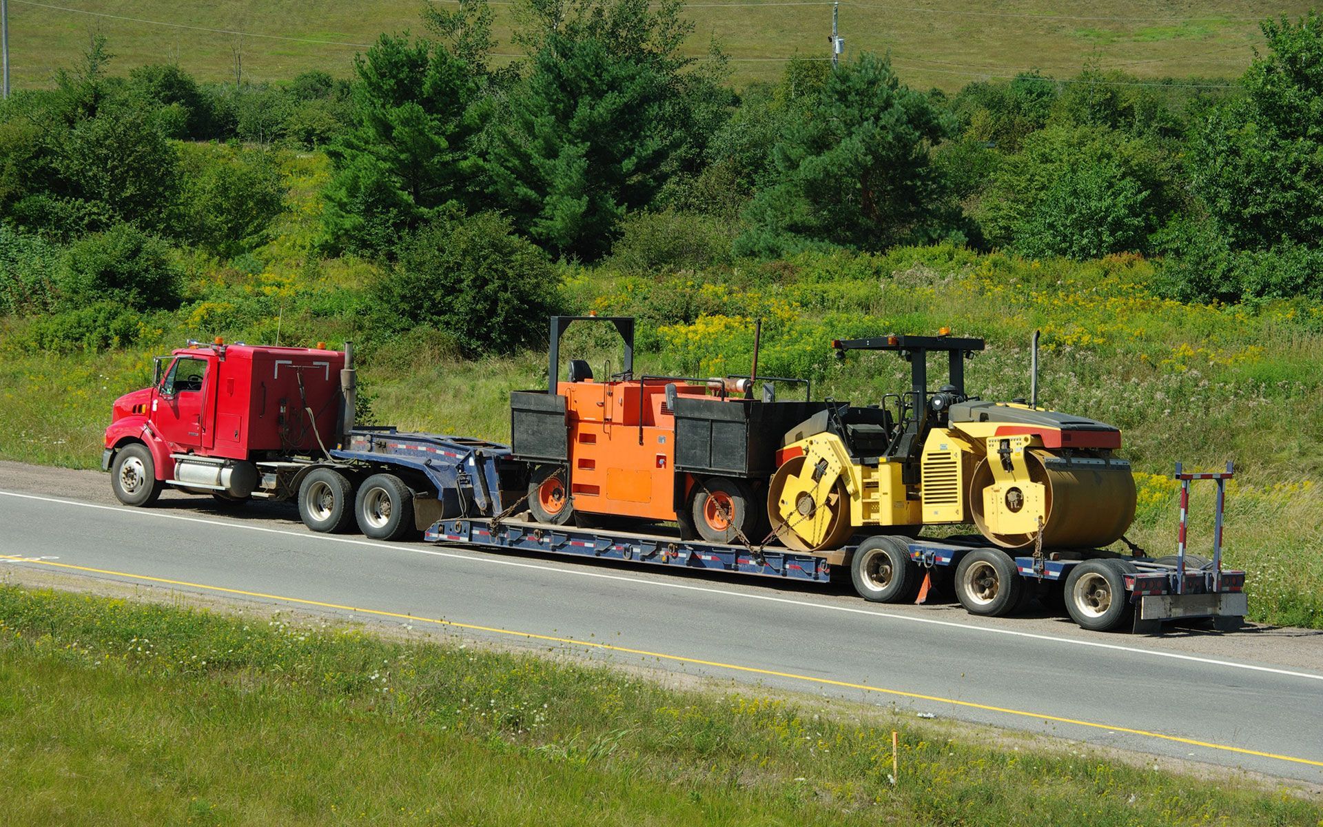 A red semi truck is driving down a highway carrying a yellow and orange tractor.