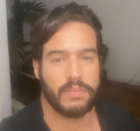 Uma pessoa com cabelo escuro e barba, vestindo uma camisa preta, olhando diretamente para a câmera.