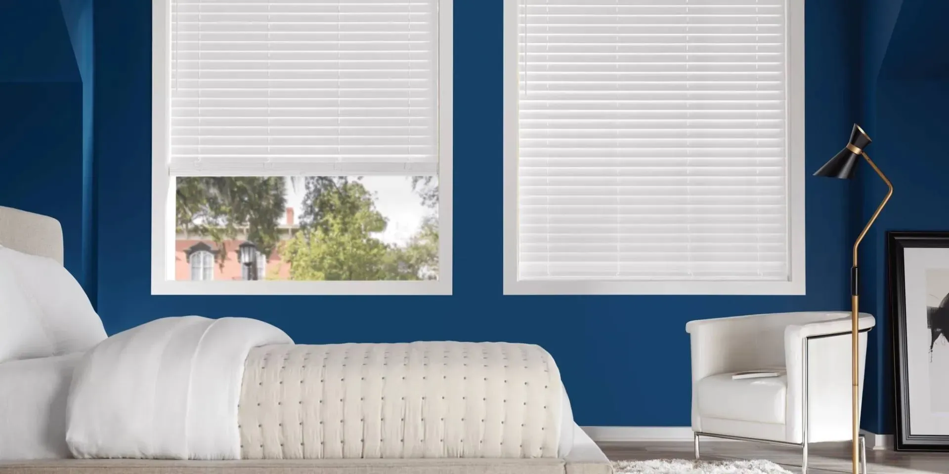 Bedroom with blue walls, white blinds, bed, chair, lamp, and framed art.