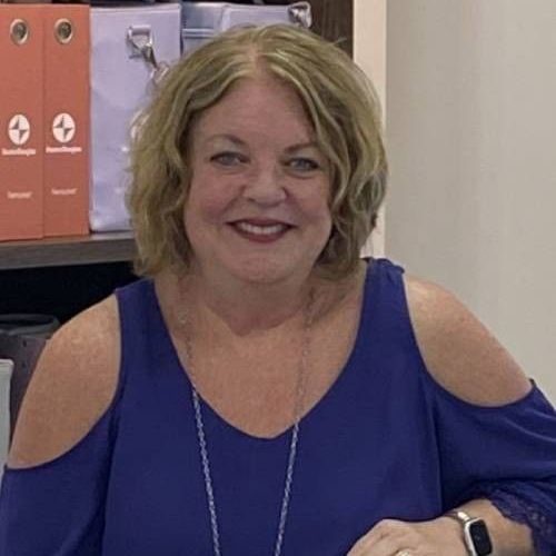 Woman with blonde hair wearing a blue, cold-shoulder top smiles, indoors.