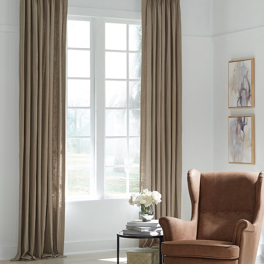 Living room with beige curtains framing a white-framed window. A brown armchair and side table with flowers complete the space.