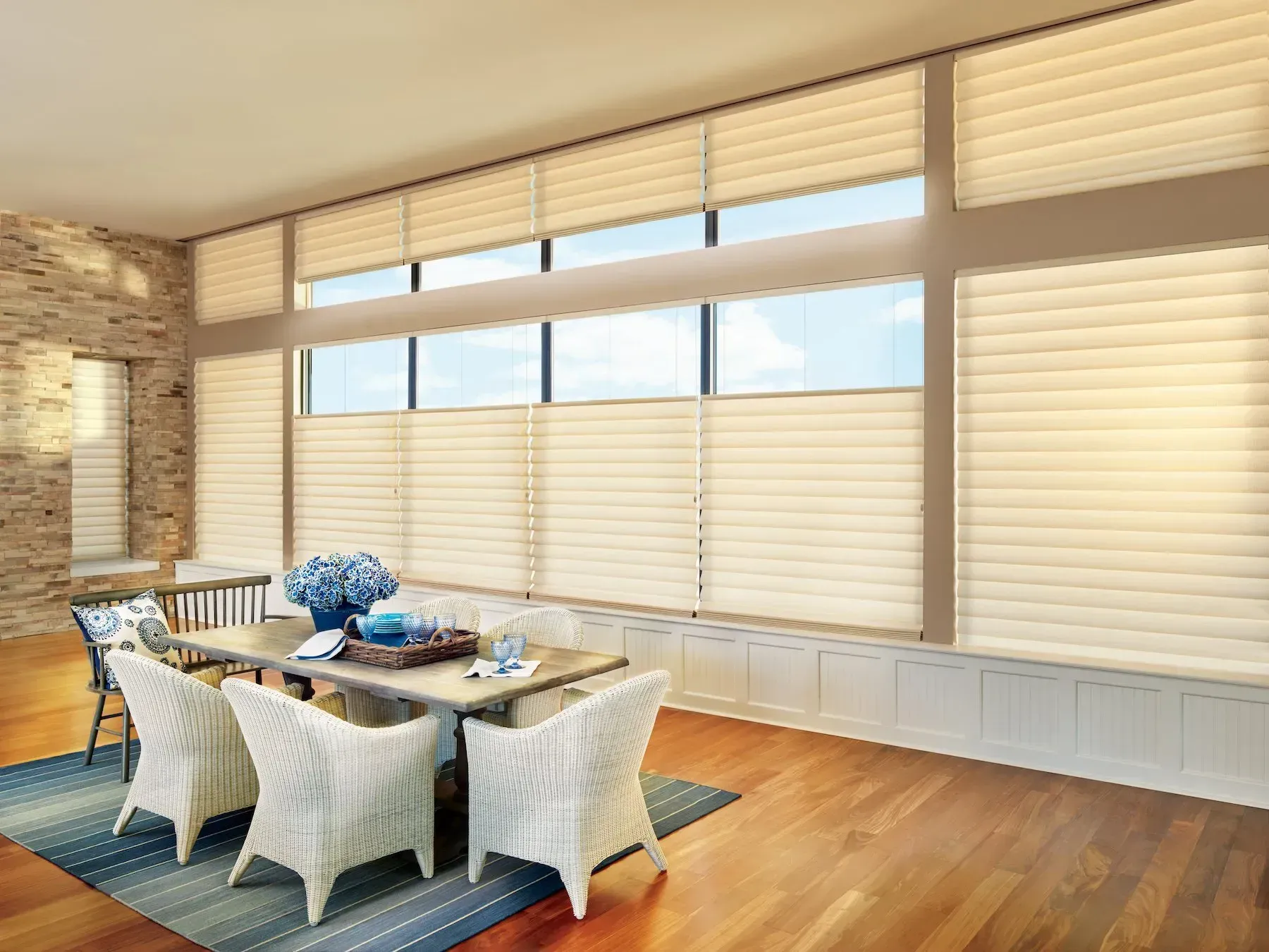 Dining room with light-colored honeycomb shades, wood floor, table with blue accents, and white wicker chairs.