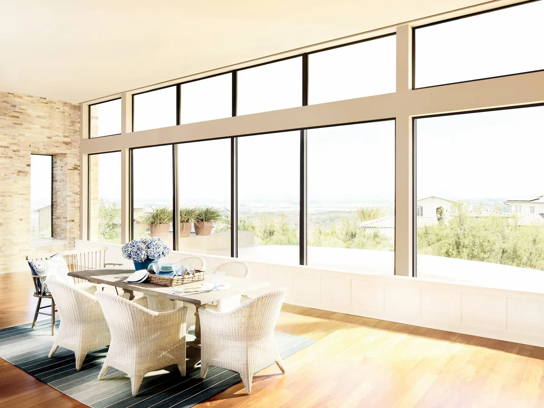 Dining room with large windows overlooking a landscape. White wicker chairs surround a table with blue accents.