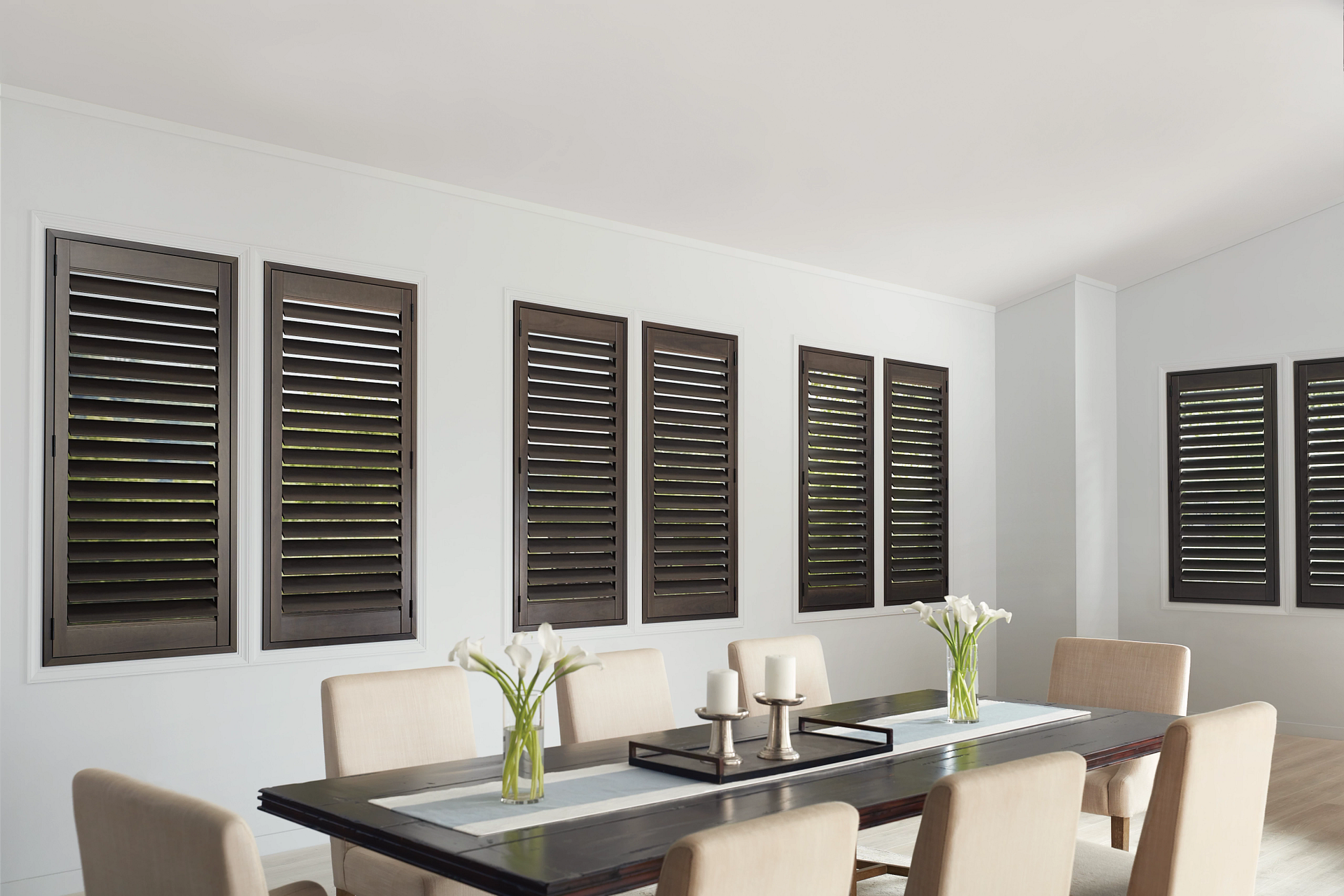 Dining room with a dark wood table, beige chairs, white walls, and dark brown shuttered windows.
