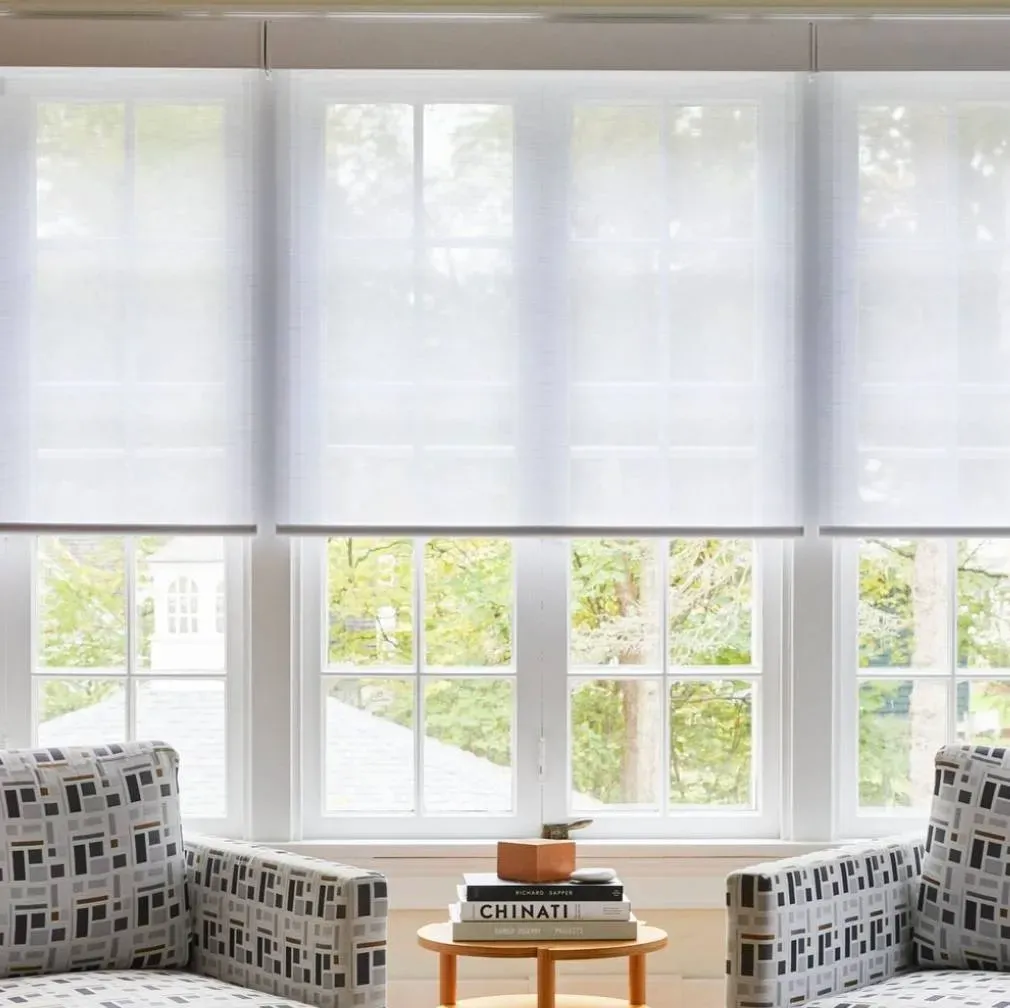 Three white roller shades covering windows; two patterned armchairs face a small table.