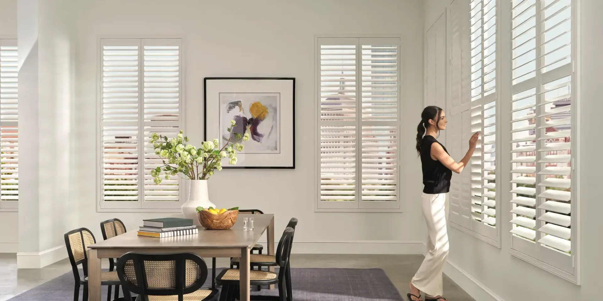 Woman adjusting white shutters in a dining room, table with fruit and floral arrangement.