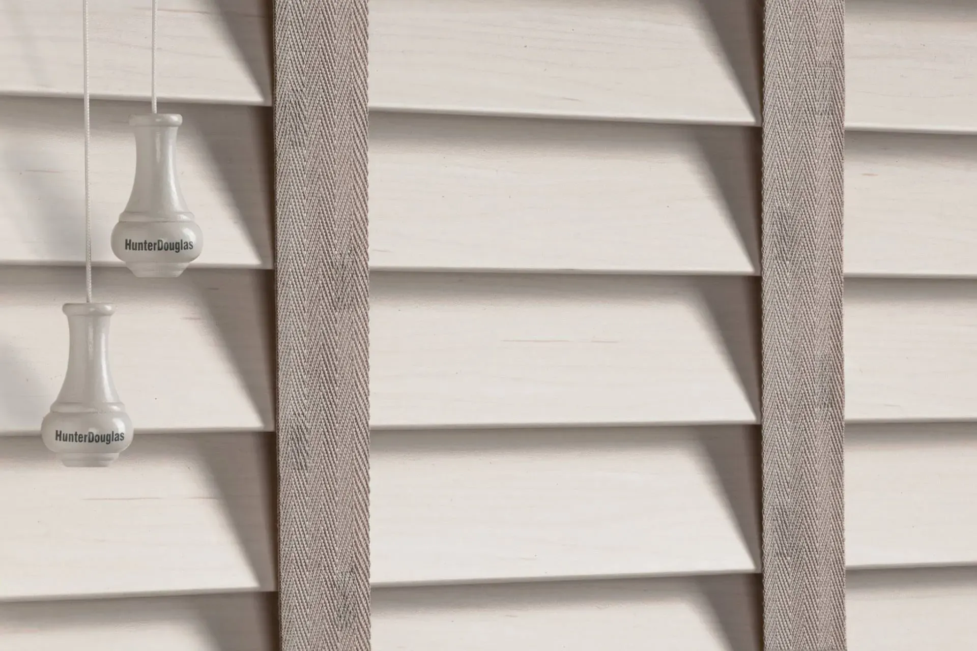 White wooden blinds with beige fabric tape and pull cords.