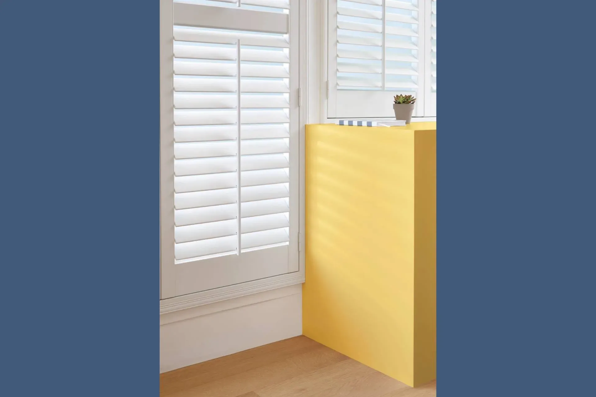 White window shutters next to a bright yellow rectangular panel, with a small plant on the ledge.