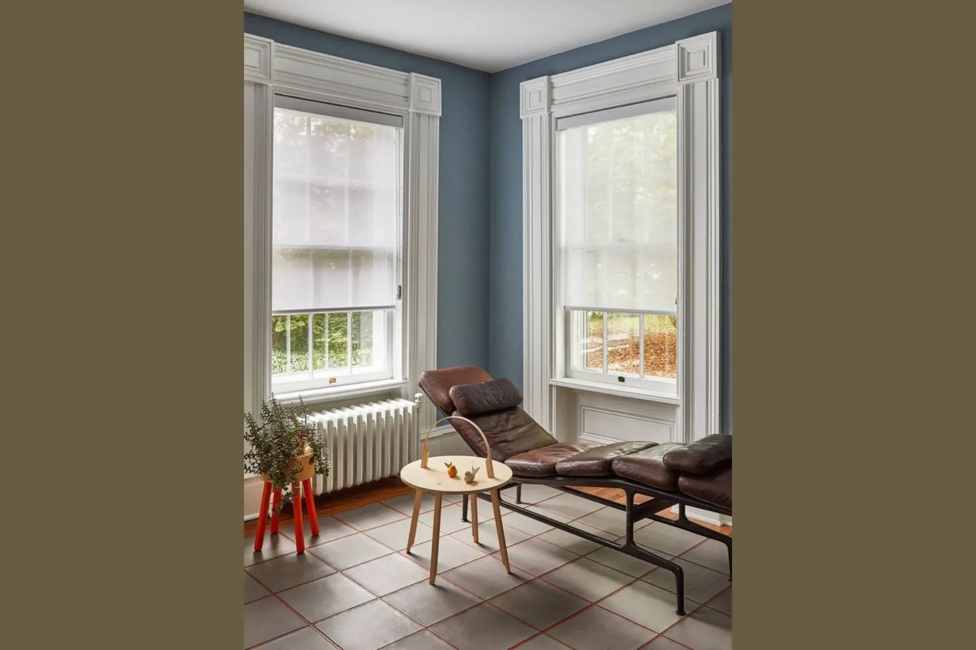 Room with blue walls, two windows with blinds, a chaise lounge, and a small table.