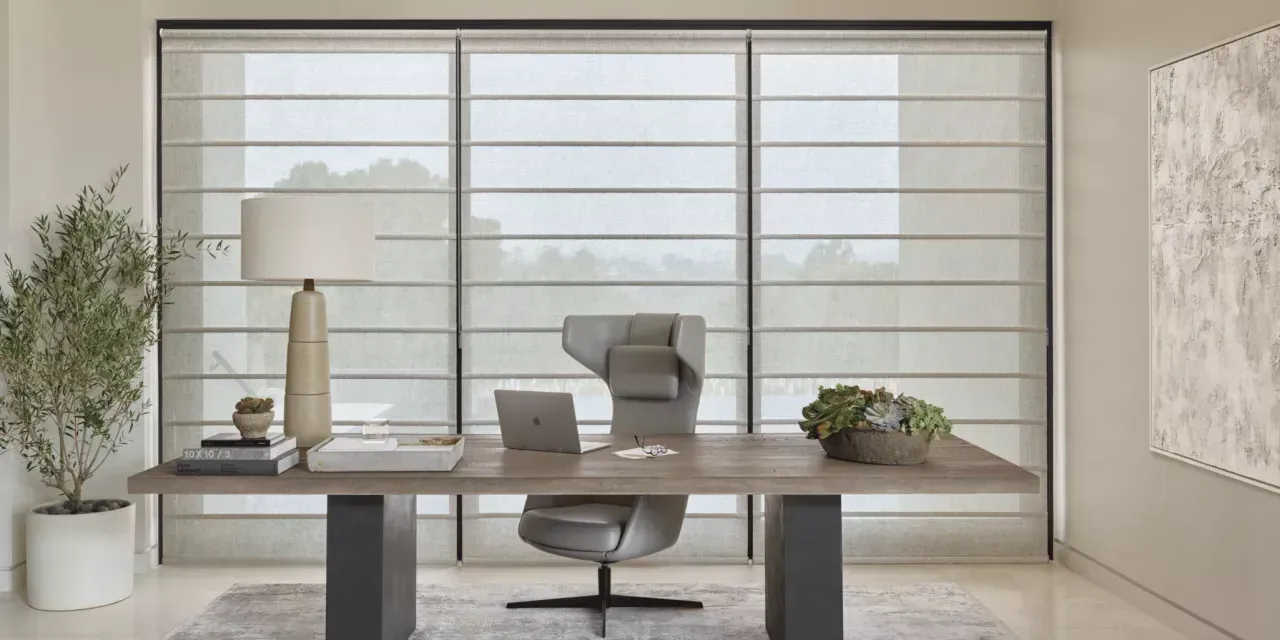 Office with desk, chair, lamp, and window with blinds. Olive tree in pot on left, artwork on right.