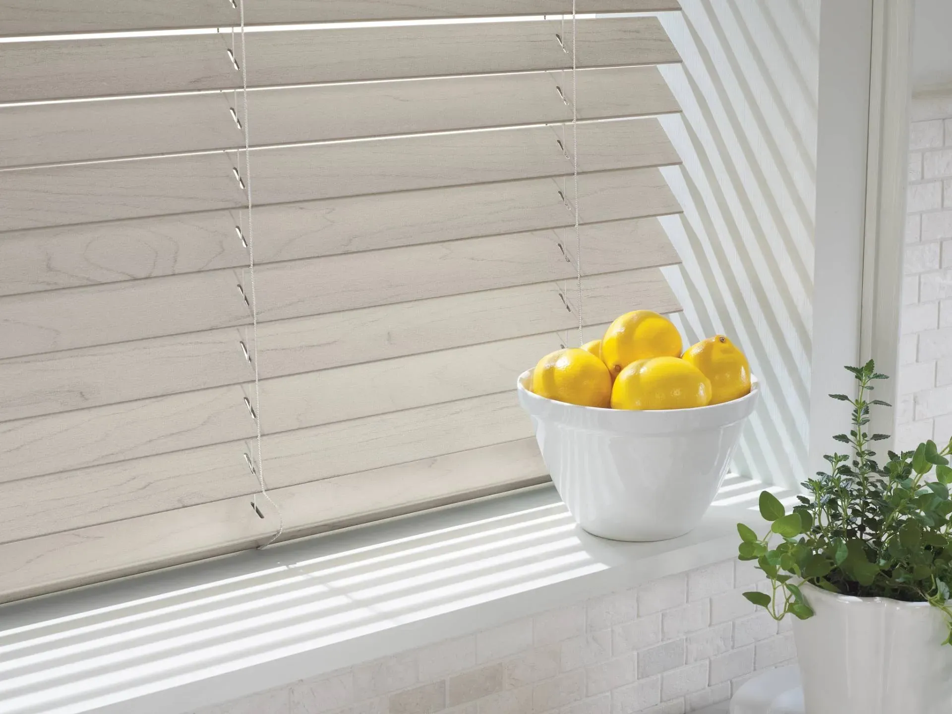 White bowl of lemons on a windowsill with closed wooden blinds casting shadows.