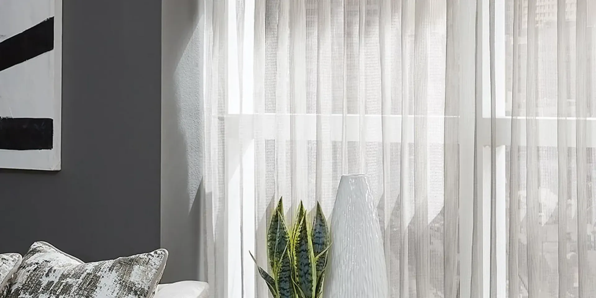White sheer curtains in front of a window, with a plant visible behind. A painting and decorative pillow are nearby.