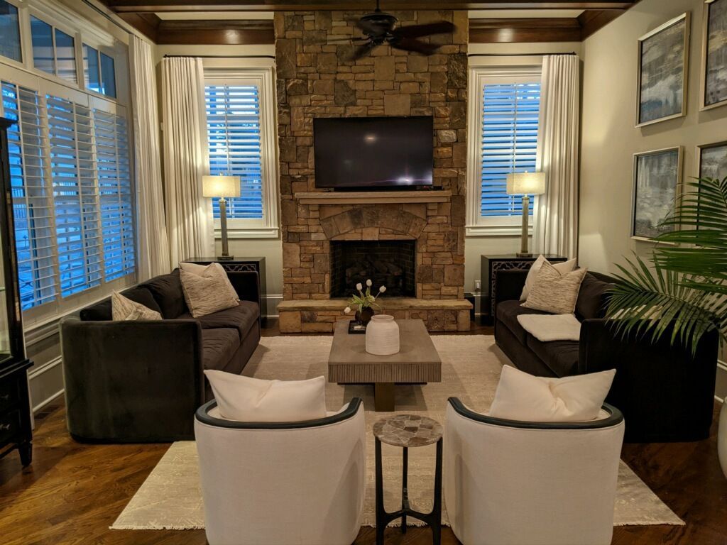 Living room with a stone fireplace, TV, two dark sofas, white chairs, and large windows.