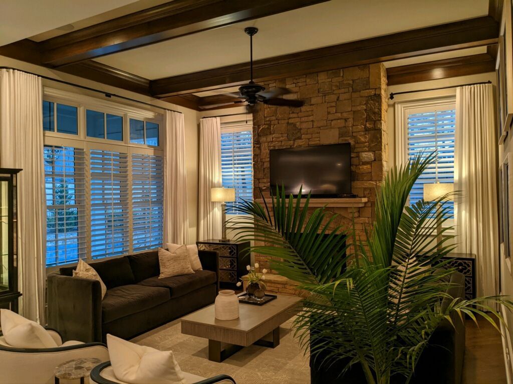 Cozy living room with stone fireplace, dark sofa, large windows with white curtains, and a ceiling fan.