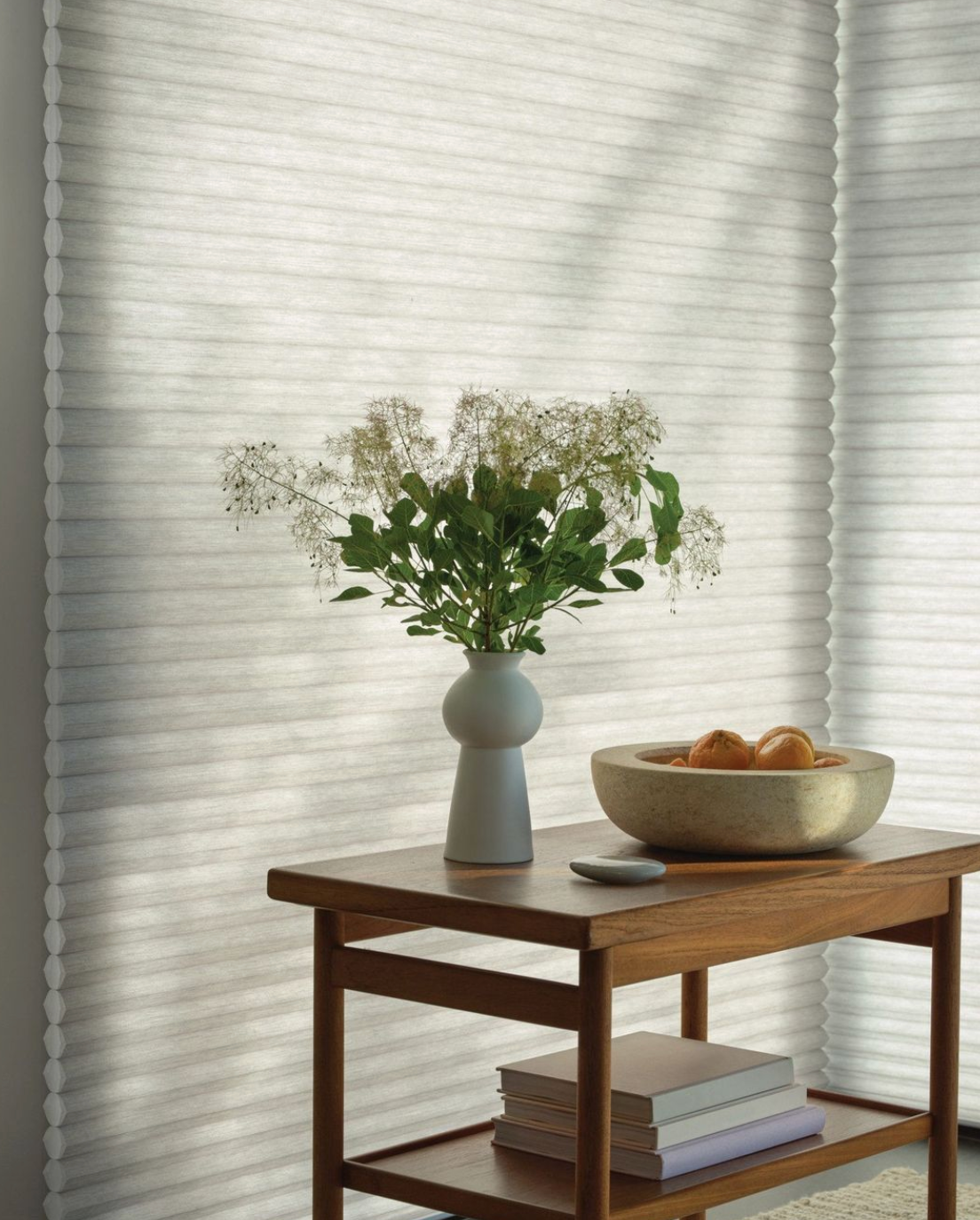Table with vase and bowl of fruit near a window with honeycomb blinds.