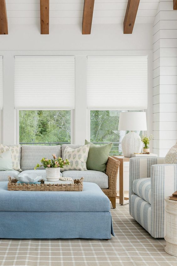 Living room with blue and neutral tones: sofa, ottoman, striped chair, white blinds, and wooden beams.
