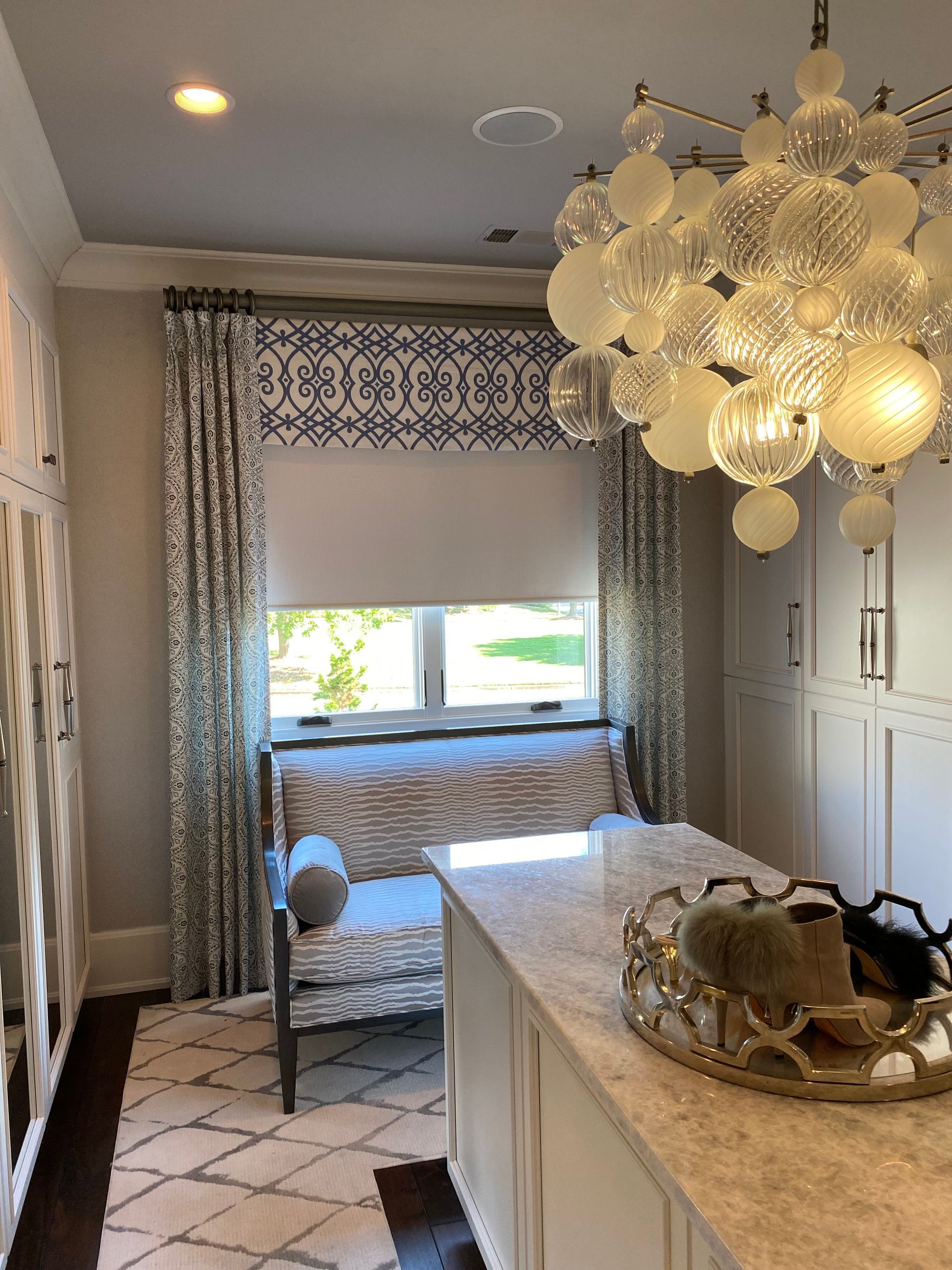 Walk-in closet with a patterned window treatment, bench, and a modern chandelier.