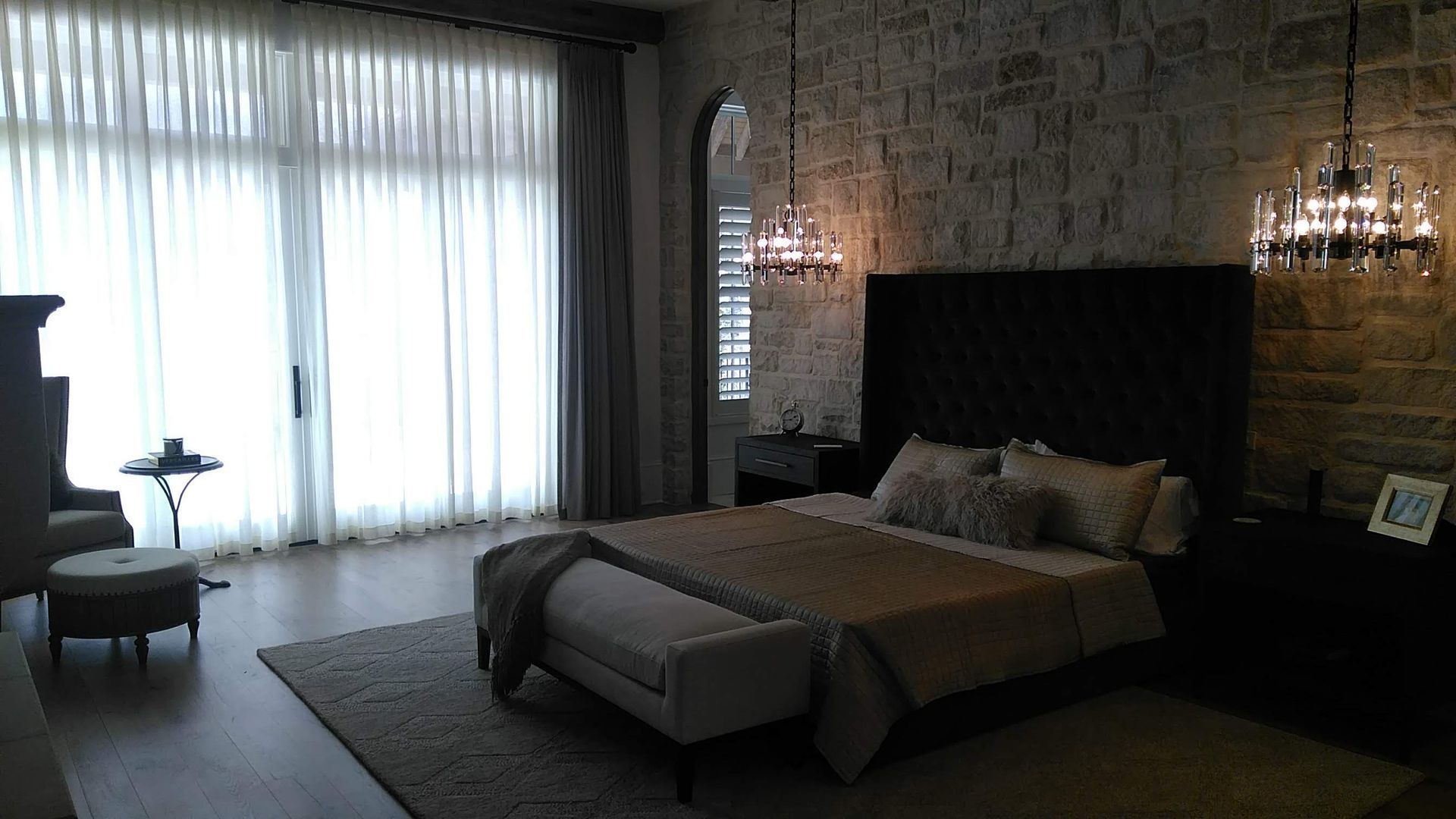 Bedroom with large bed, stone wall, chandeliers, sheer curtains over a large window, and wooden floors.