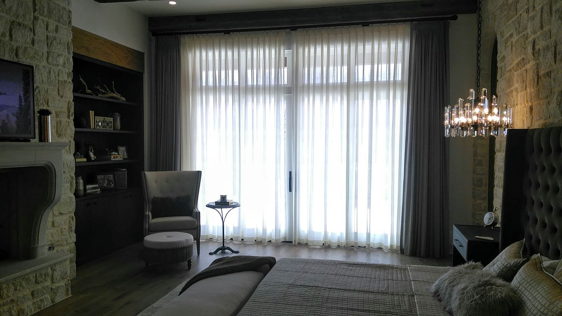 Bedroom with sheer curtains, stone walls, chandelier, armchair, and built-in shelves.