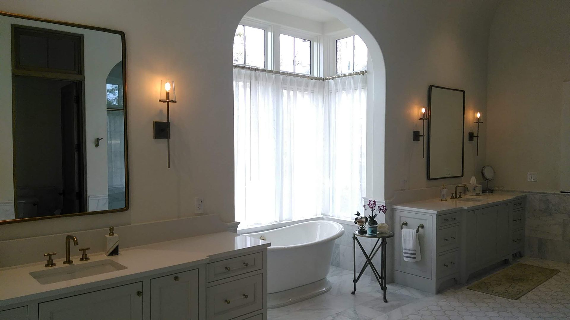 Bathroom with arched window, white walls, freestanding tub, two vanities, and decorative lighting.