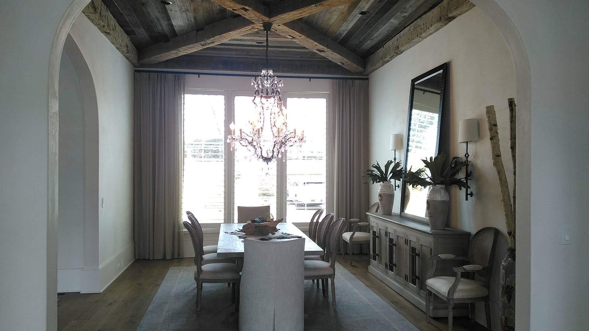Dining room with long table, chandelier, and tall windows; neutral colors, rustic ceiling.