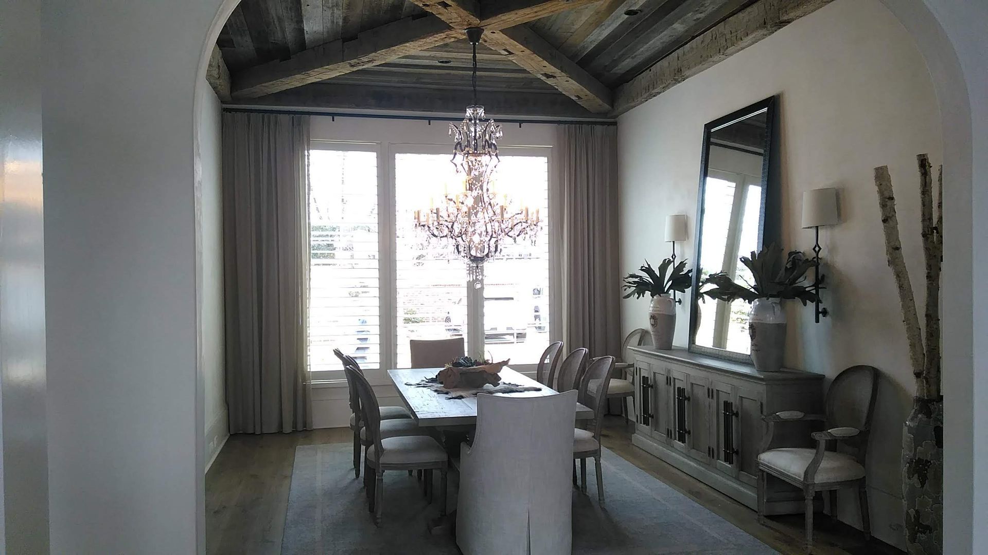 Dining room with wooden ceiling, chandelier, long table, buffet, and large mirror.