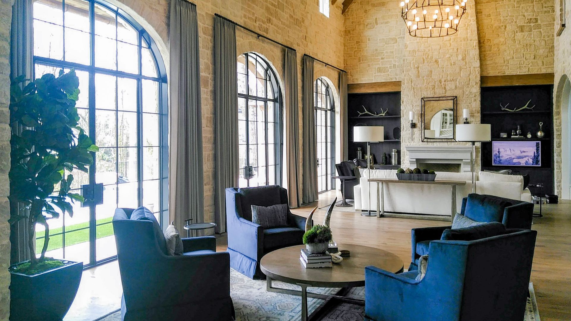 Living room with blue armchairs, round coffee table, arched windows, stone walls, and chandelier.