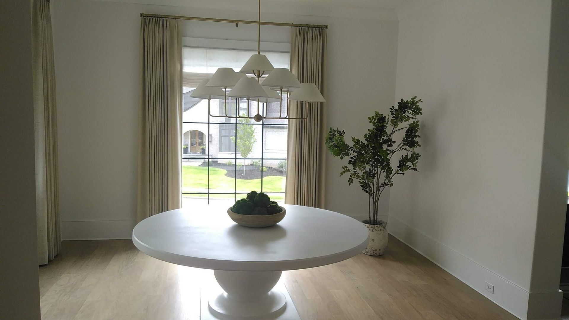Round white table with greenery, under a chandelier near a window with curtains and a potted plant.