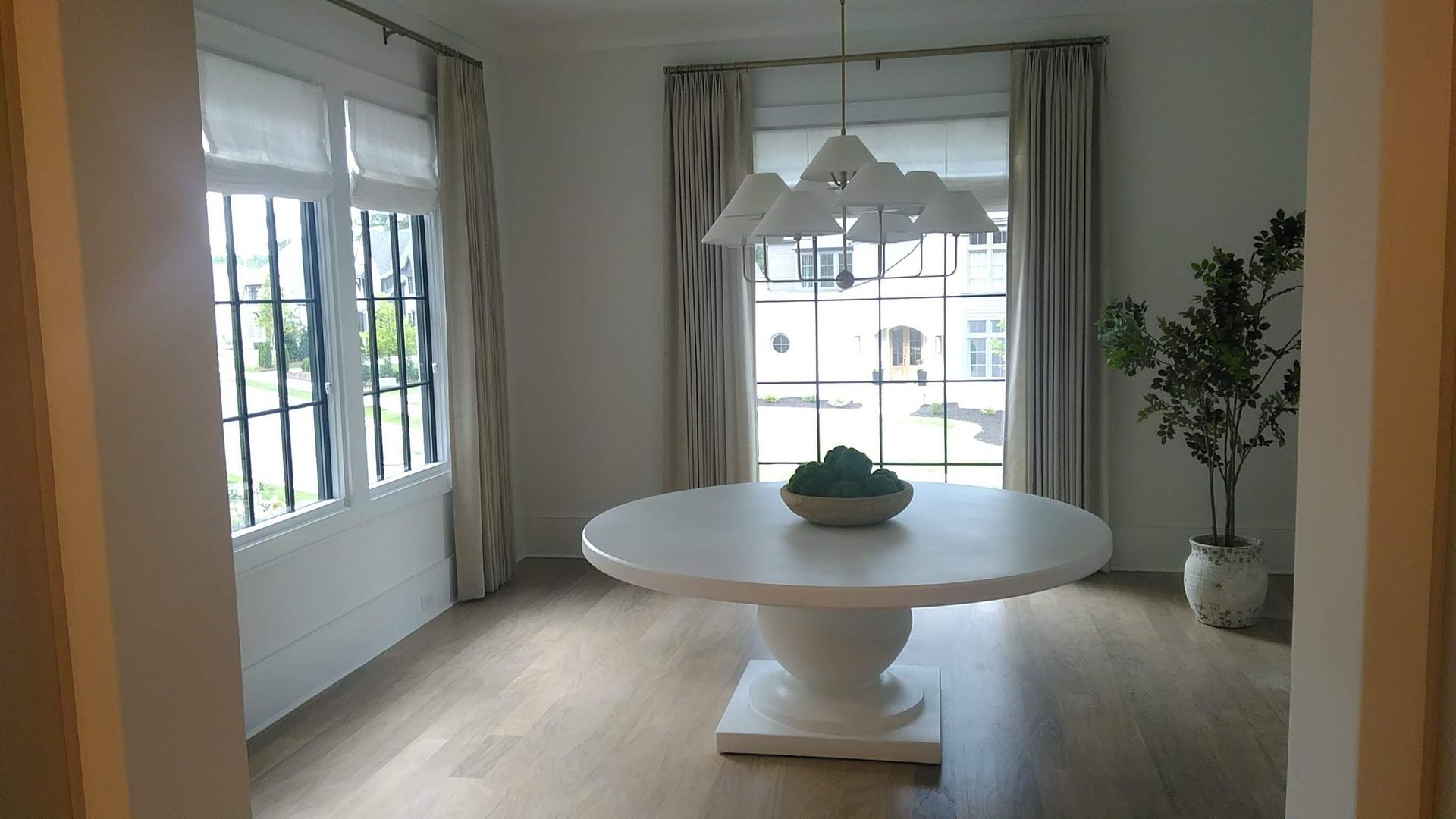 Round white table in a light-filled dining room with a chandelier, curtains, and an indoor tree.