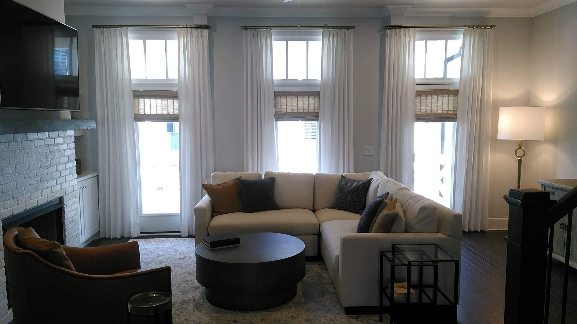 Living room with white sectional sofa, three windows with white curtains and woven shades.