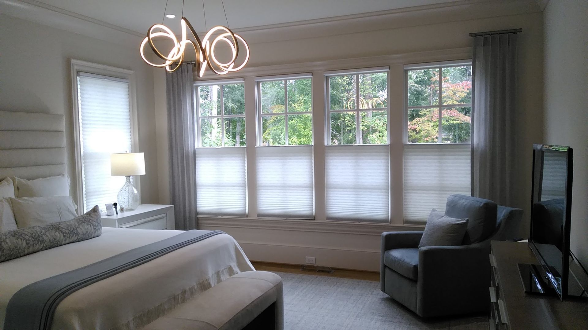 Bedroom with bed, chair, windows with light filtering shades and drapes, and a chandelier.