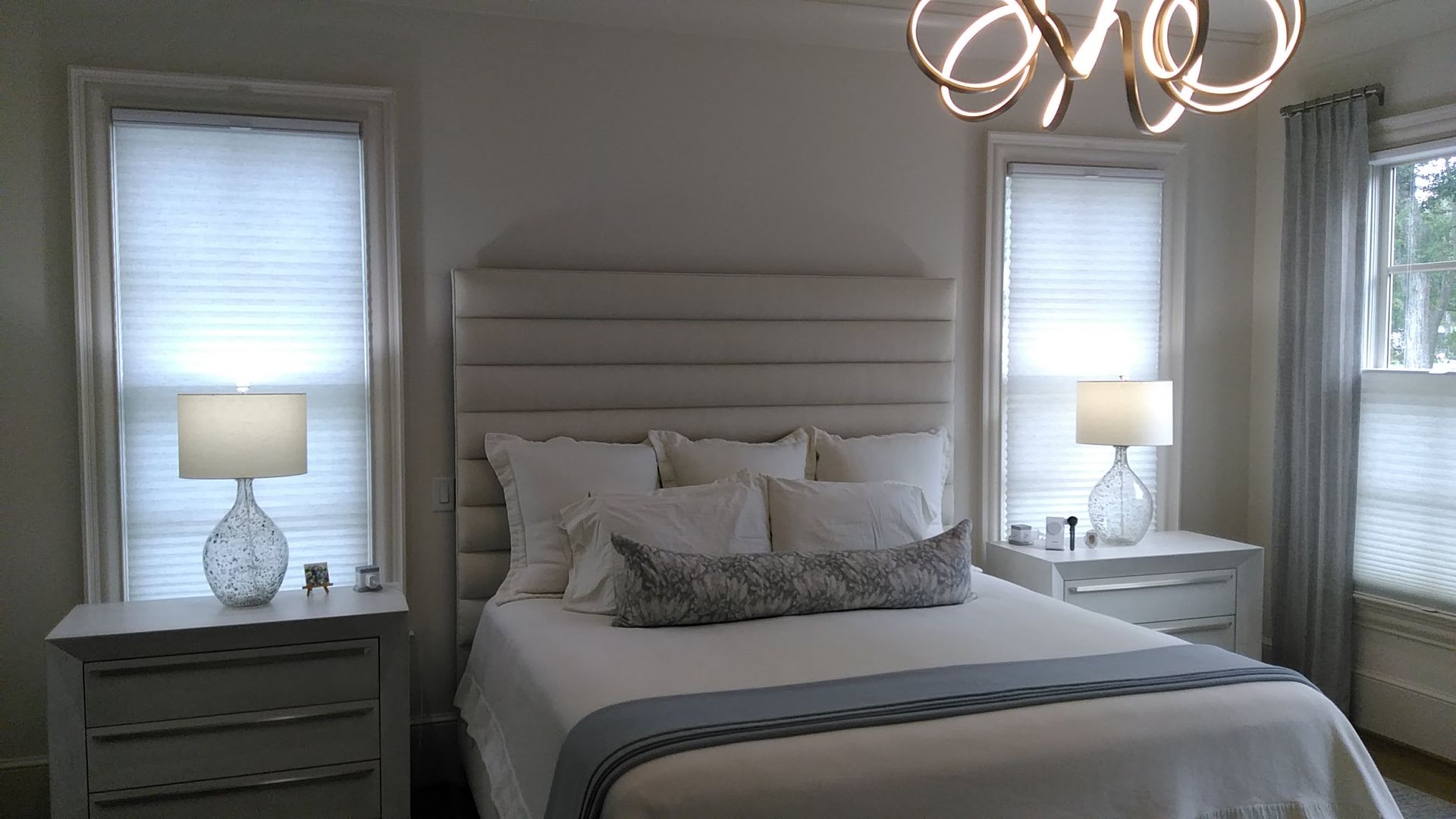 Bedroom with white bed, upholstered headboard, two nightstands, two windows with blinds.