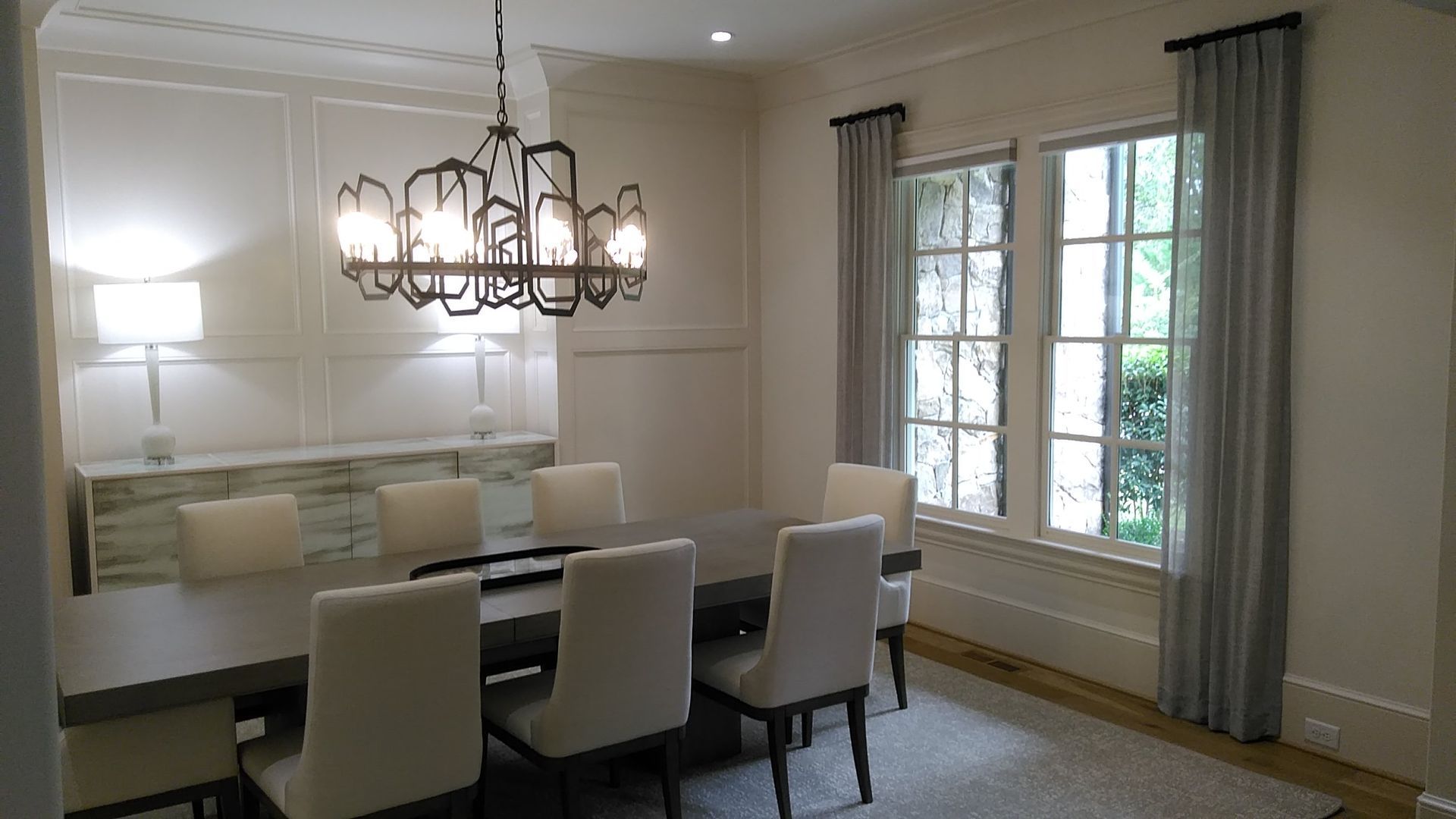 Dining room with a long table, chandelier, chairs, windows with curtains.