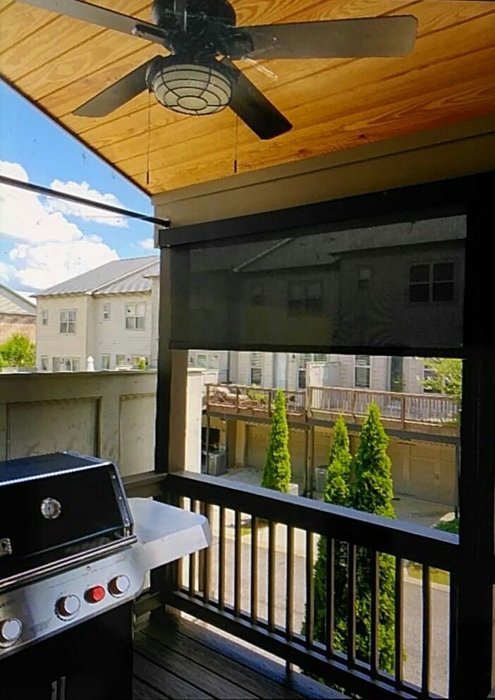 Patio with a grill, black sunshade, and ceiling fan. View of buildings and trees.