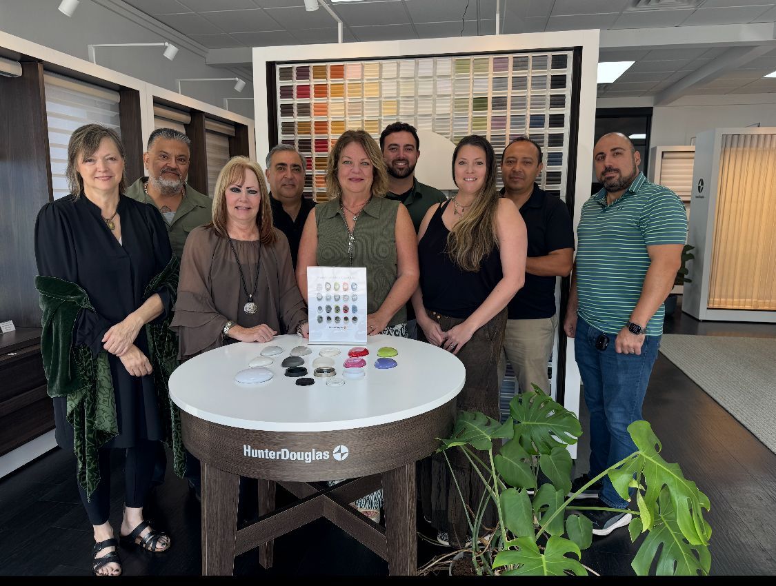 Group of people smiling, posing around a display table in a store with color samples and window treatments in the background.