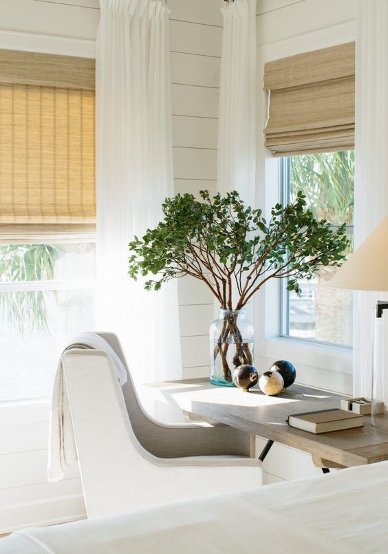 Bright home office with desk, chair, and large tree in a vase by window, with light curtains and blinds.