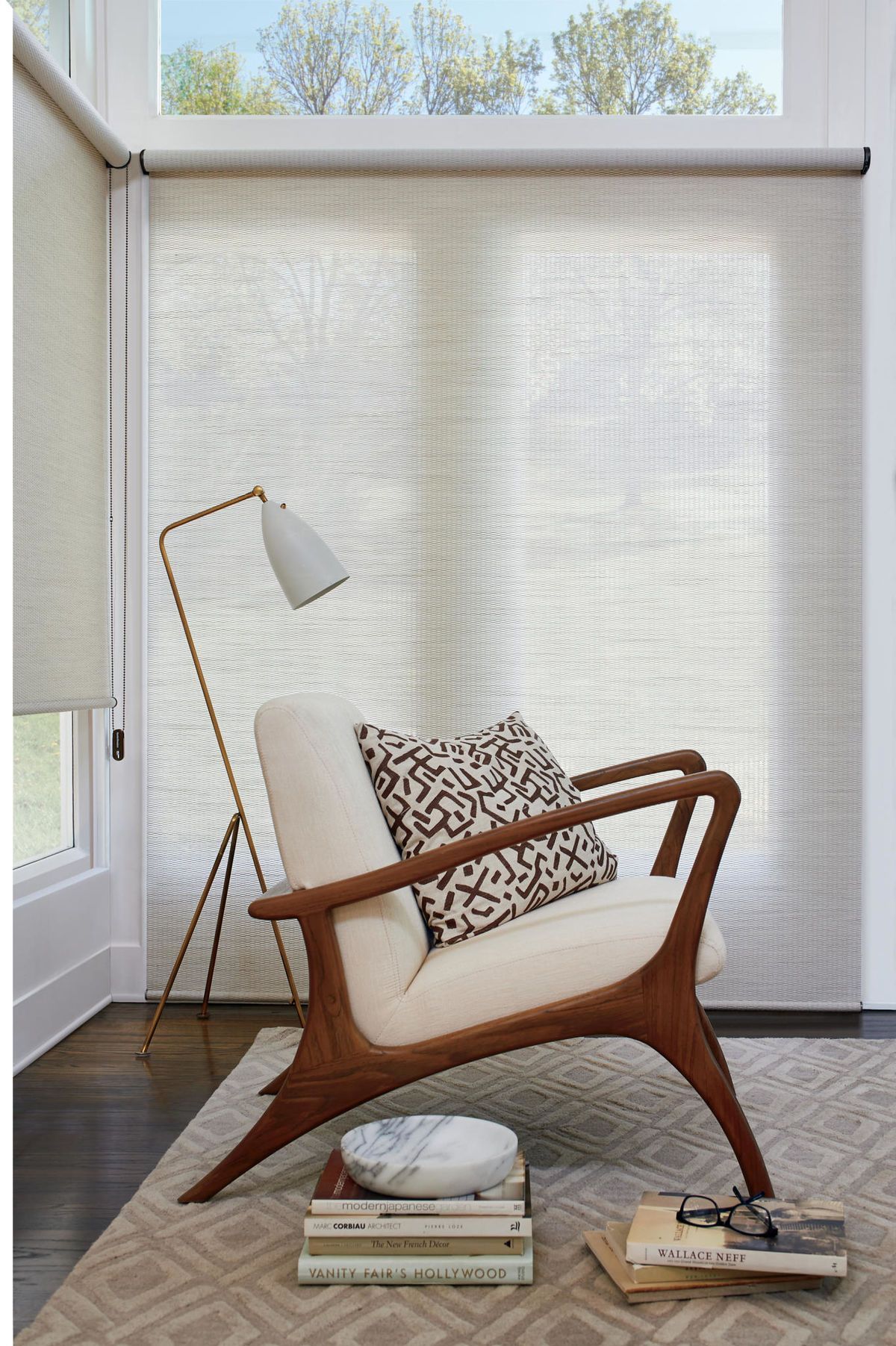 Stylish chair with a pillow, floor lamp, books, and woven blinds in a sunlit room.