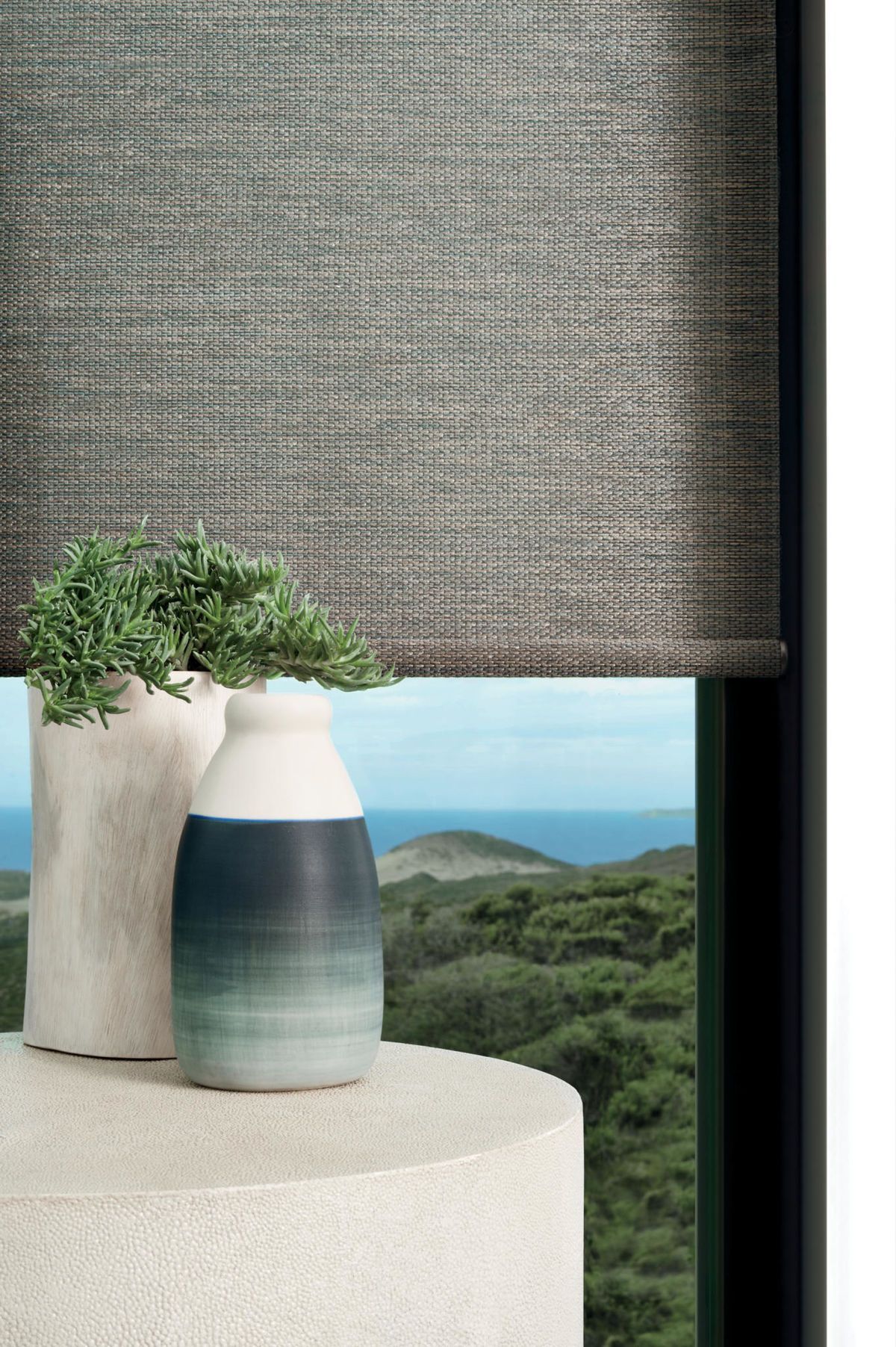 Woven textured roller shade over a window with two vases and greenery. Ocean and hills in background.
