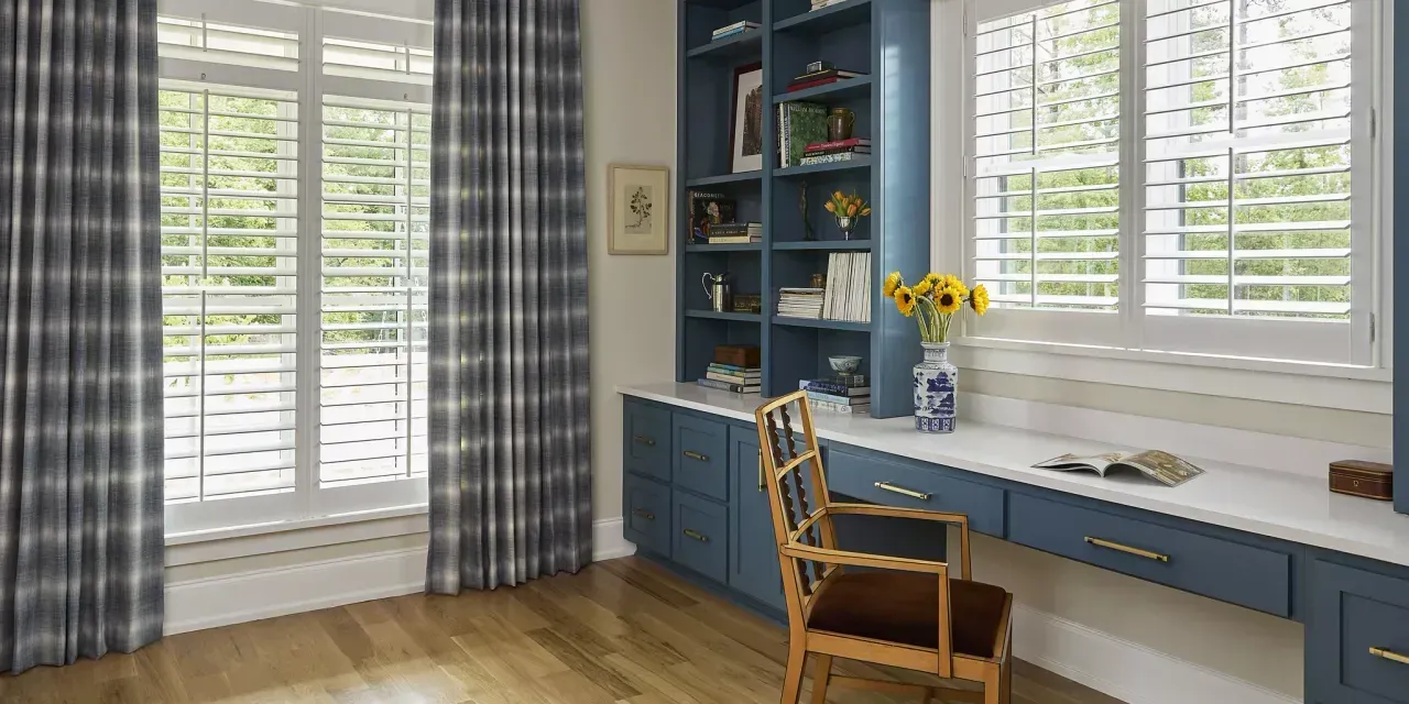Home office with blue built-in desk and bookshelves, window with shutters and plaid curtains, wooden floor.
