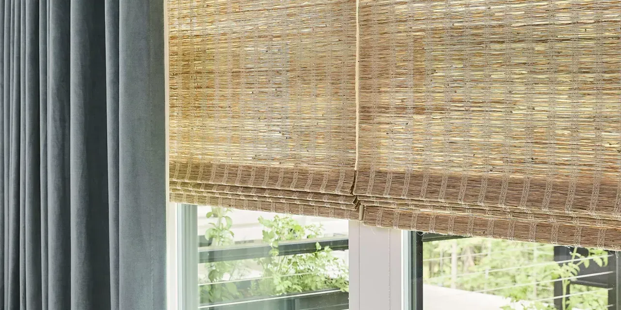Window with woven shade and blue curtains, overlooking greenery.