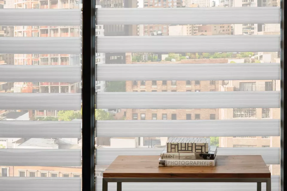 Window with layered white and sheer blinds overlooking city buildings. Wooden table holds books.