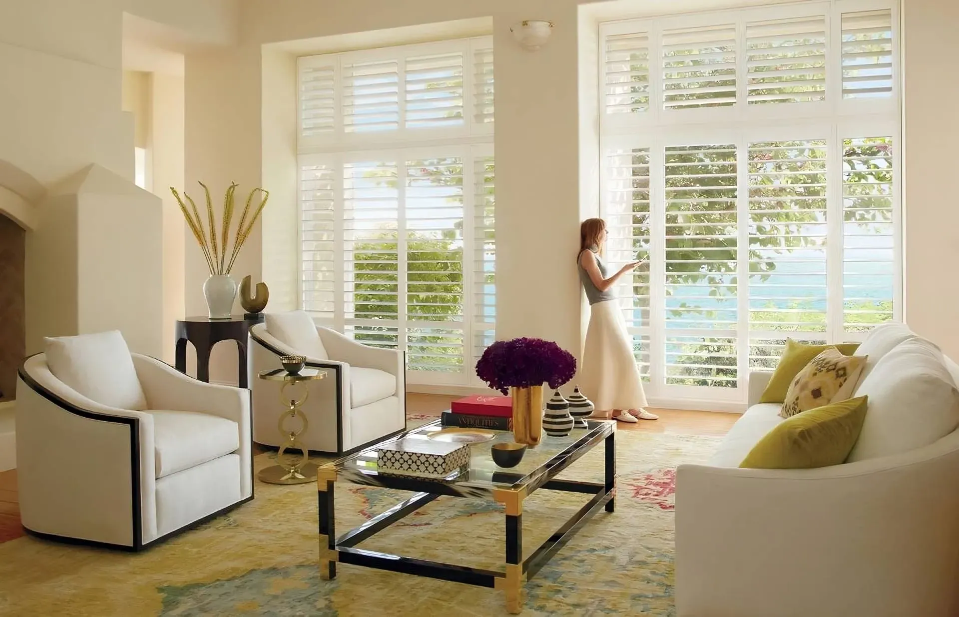 Living room with white shutters, a woman looking out, and white furniture.