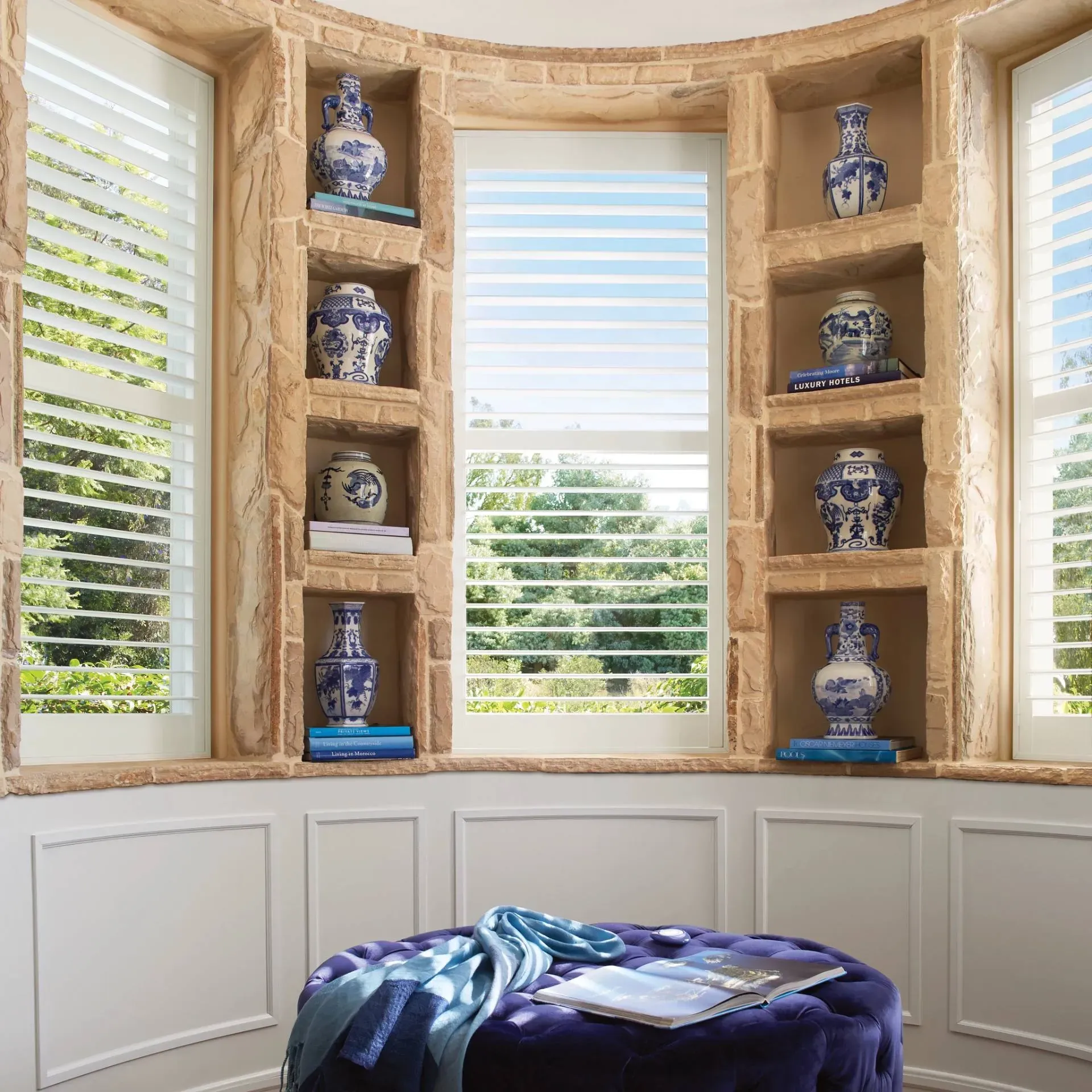 Bay window with built-in shelves displaying blue and white vases. The window has shutters and overlooks a green landscape.