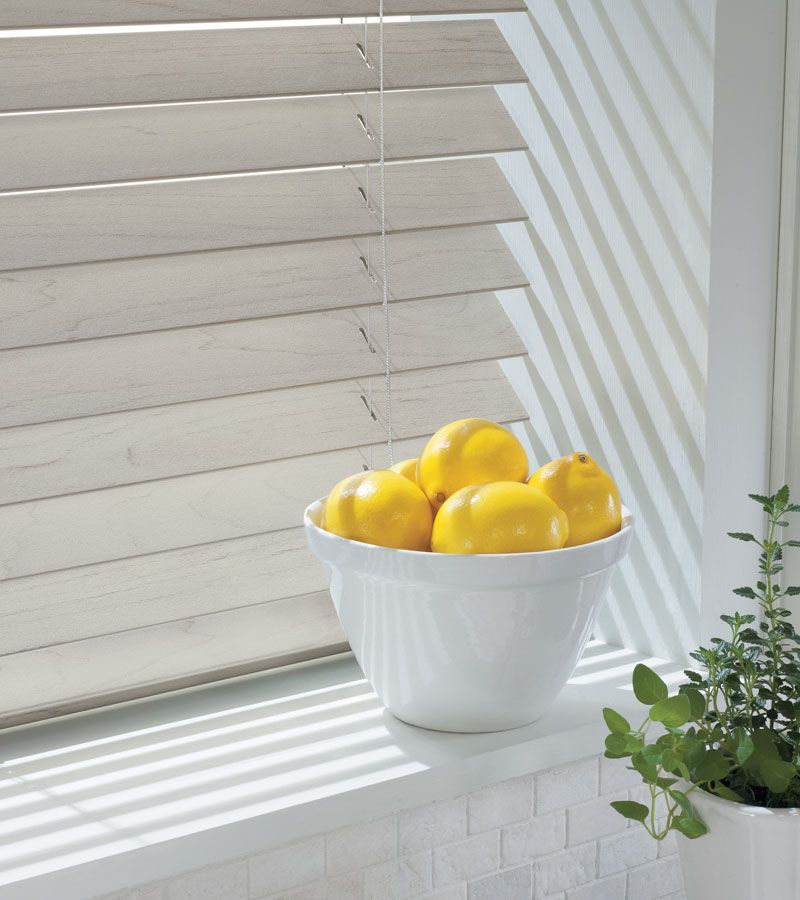 A white bowl of lemons sits on a windowsill with wooden blinds and sunlight.
