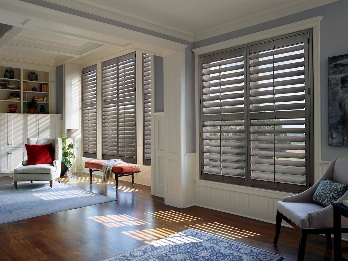 Living room with gray shutters, hardwood floors, blue rug, and seating. Sunlight streams through.