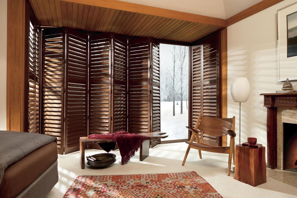 Wooden shutters open to snowy outdoors in a cozy room, with seating, rug, and fireplace.